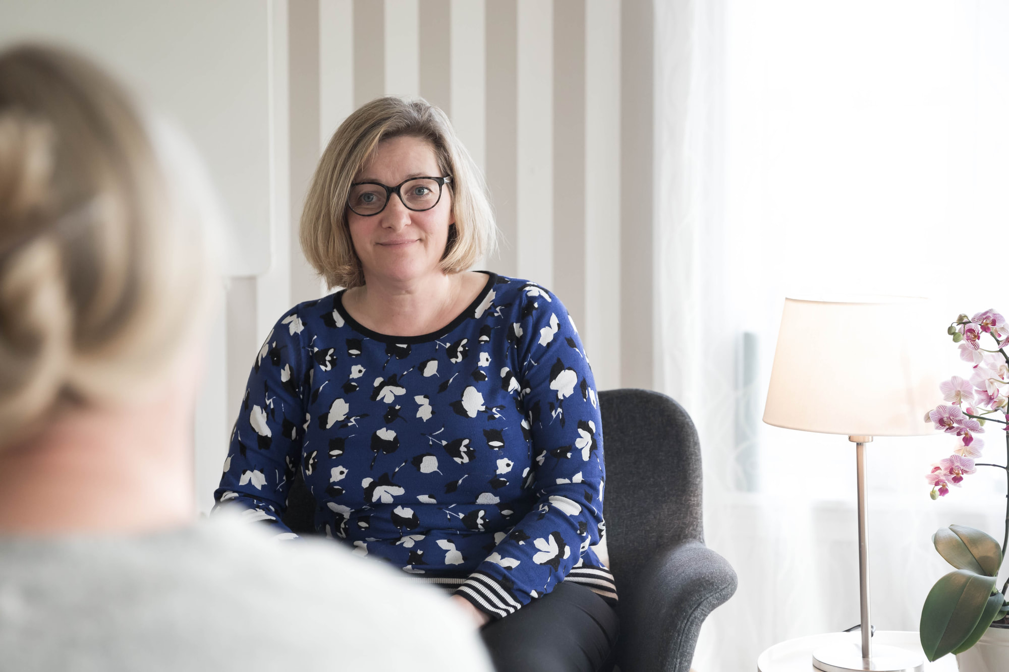 Lotte sidder i samtale med en klient i terapihuset, med en tændt lampe og en orkidé i baggrunden